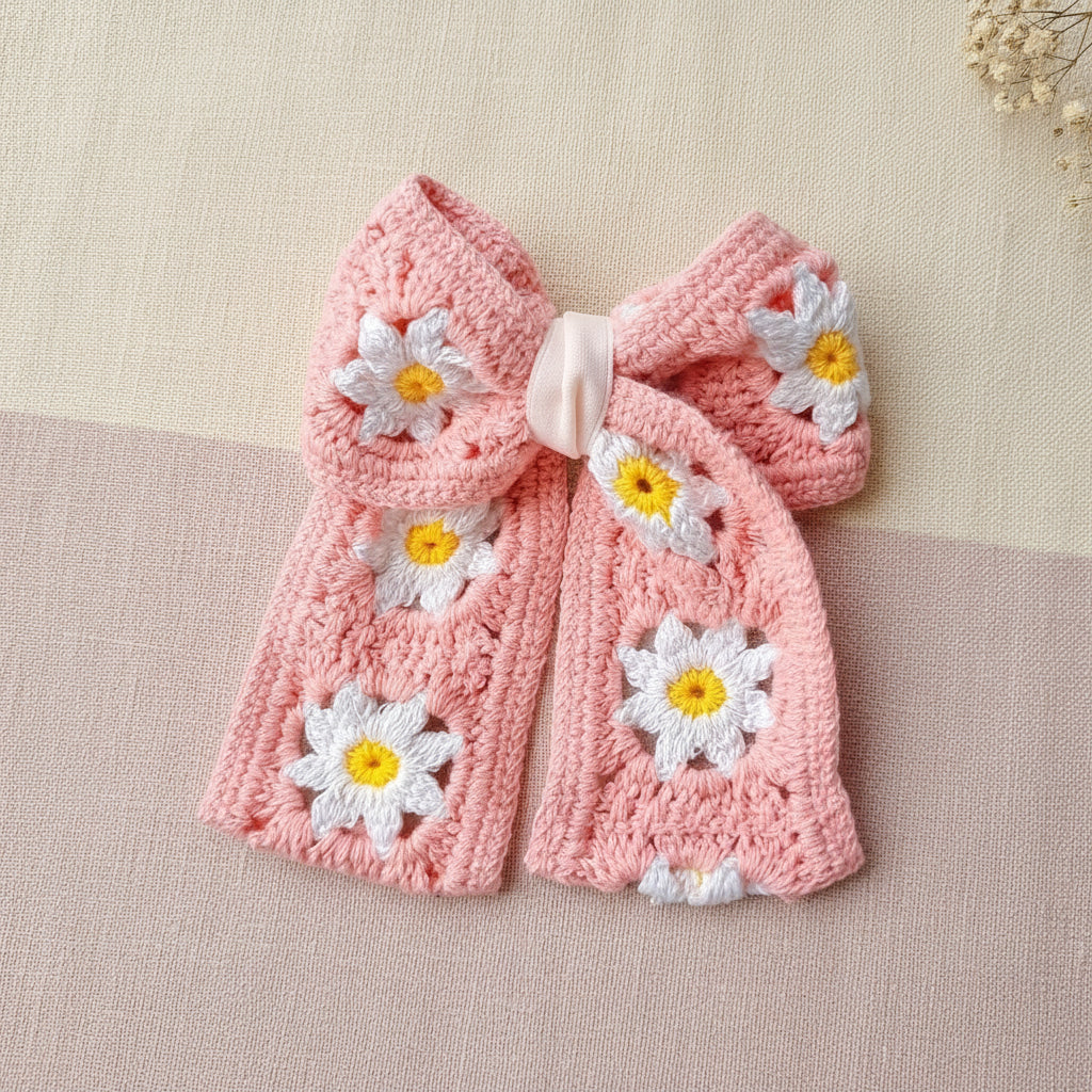 A pink crocheted pet collar with a bow and daisy flower details, laid flat on a light-colored surface.