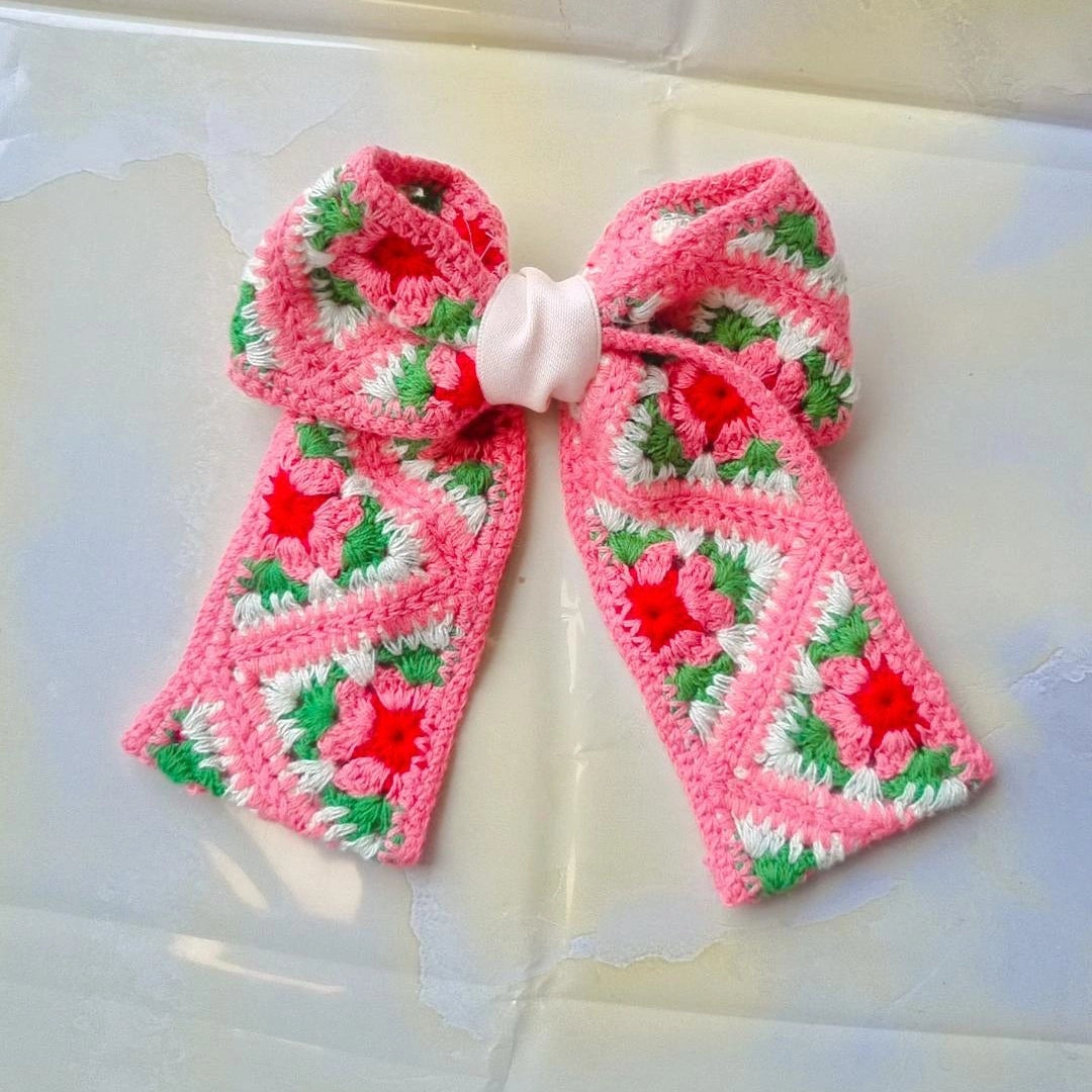A crocheted hair bow featuring a pink and white granny square pattern with red and green floral accents, tied with a white ribbon at the center, displayed on a light-colored surface.