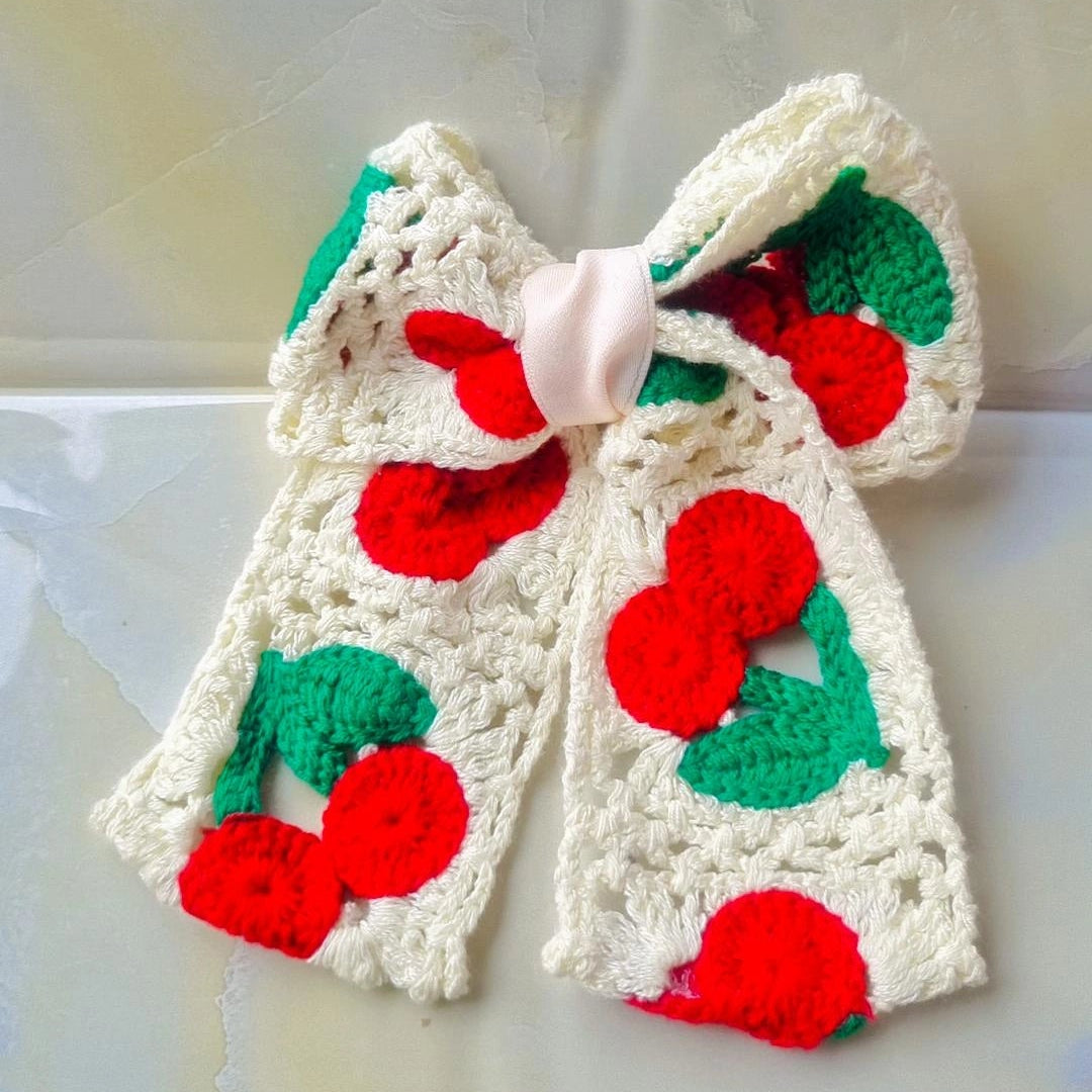 A handmade crocheted hair bow featuring a cherry pattern with red yarn cherries and green yarn leaves on an off-white base, finished with a small pale pink center ribbon.