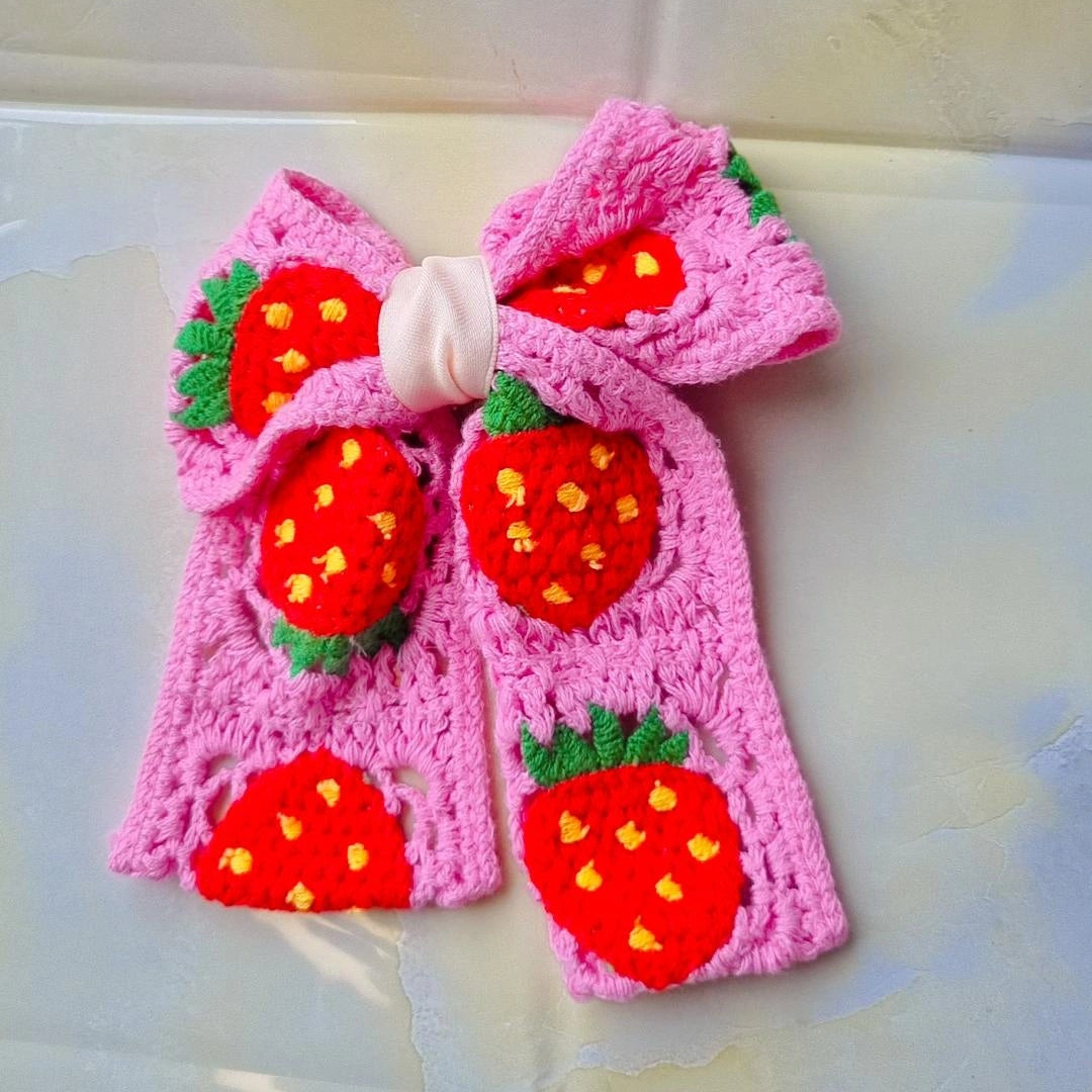 A pink crocheted bow headband decorated with three red and yellow strawberry appliqués and green leafy tops, displayed on a light-colored surface.