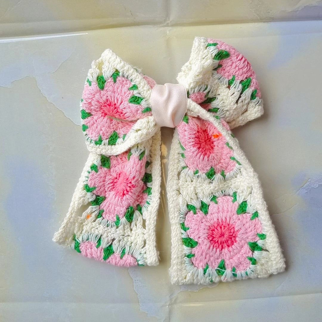 A handmade crochet bow headband featuring pink floral patterns with green leaves on an off-white base, laid flat on a light-colored tiled surface.
