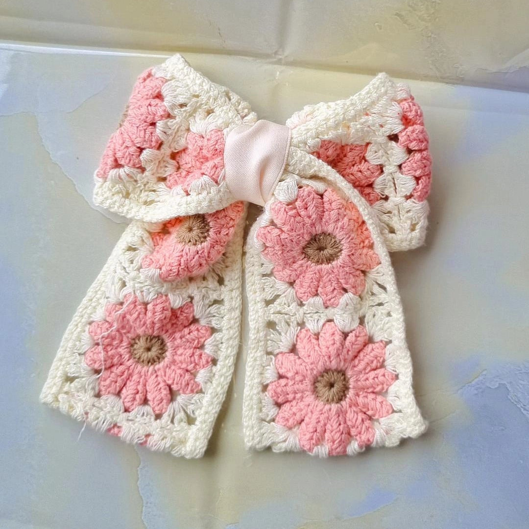 A handmade crocheted hair bow accessory featuring pink and cream crochet floral motifs and a light pink satin center ribbon, displayed on a light-colored surface.