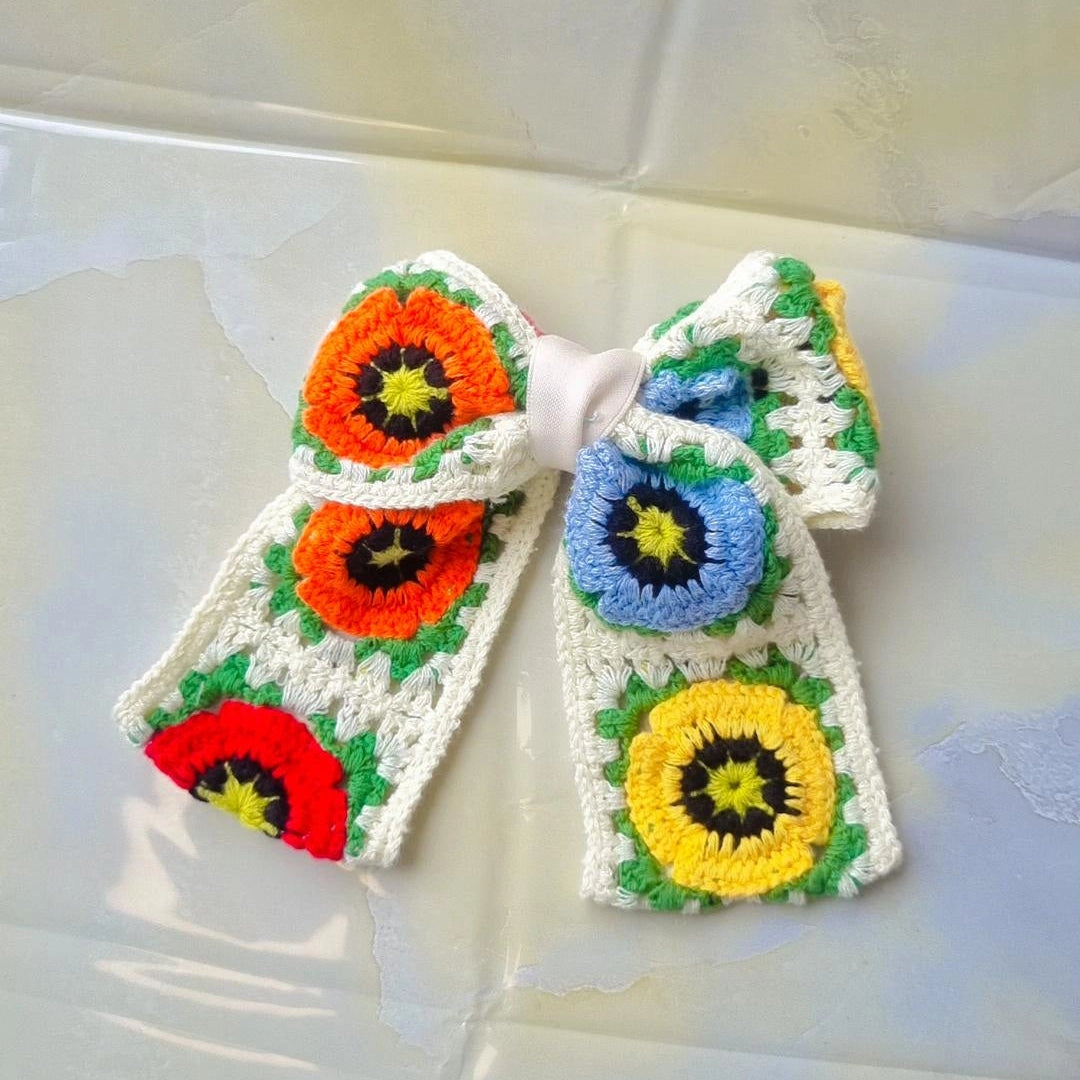 A handmade crocheted hair bow featuring colorful floral motifs in orange, red, blue, yellow, and green on a white crochet base, displayed on a glossy, reflective light-colored surface.