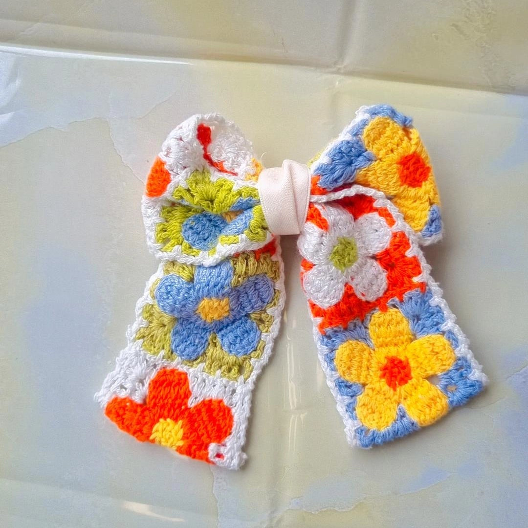 A handmade crocheted bow hair clip featuring a colorful floral pattern with blue, yellow, orange, and white flowers on a white background, secured with a white plastic pin.