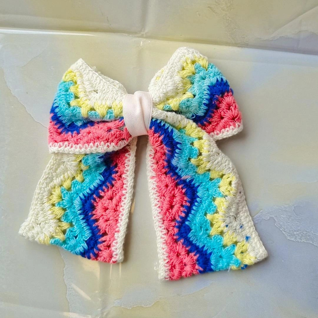 A handmade crochet hair bow with a colorful zigzag pattern in pink, blue, yellow, and green on a white base, tied with a white ribbon at the center, resting on a glossy, light-colored surface.