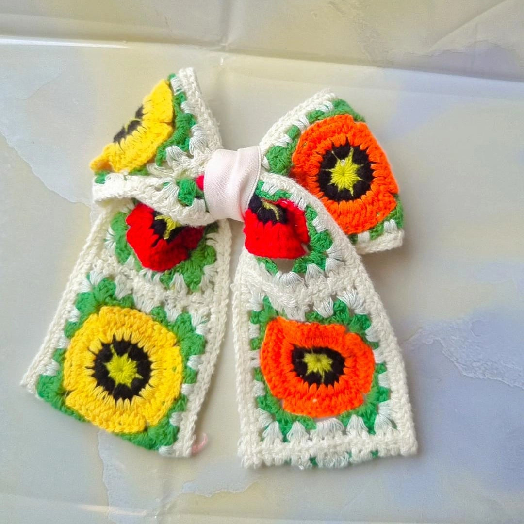 A handmade crocheted hair bow featuring colorful sunflower and poppy motifs in yellow, orange, red, and green on a white crochet base, tied in the center with a white ribbon.