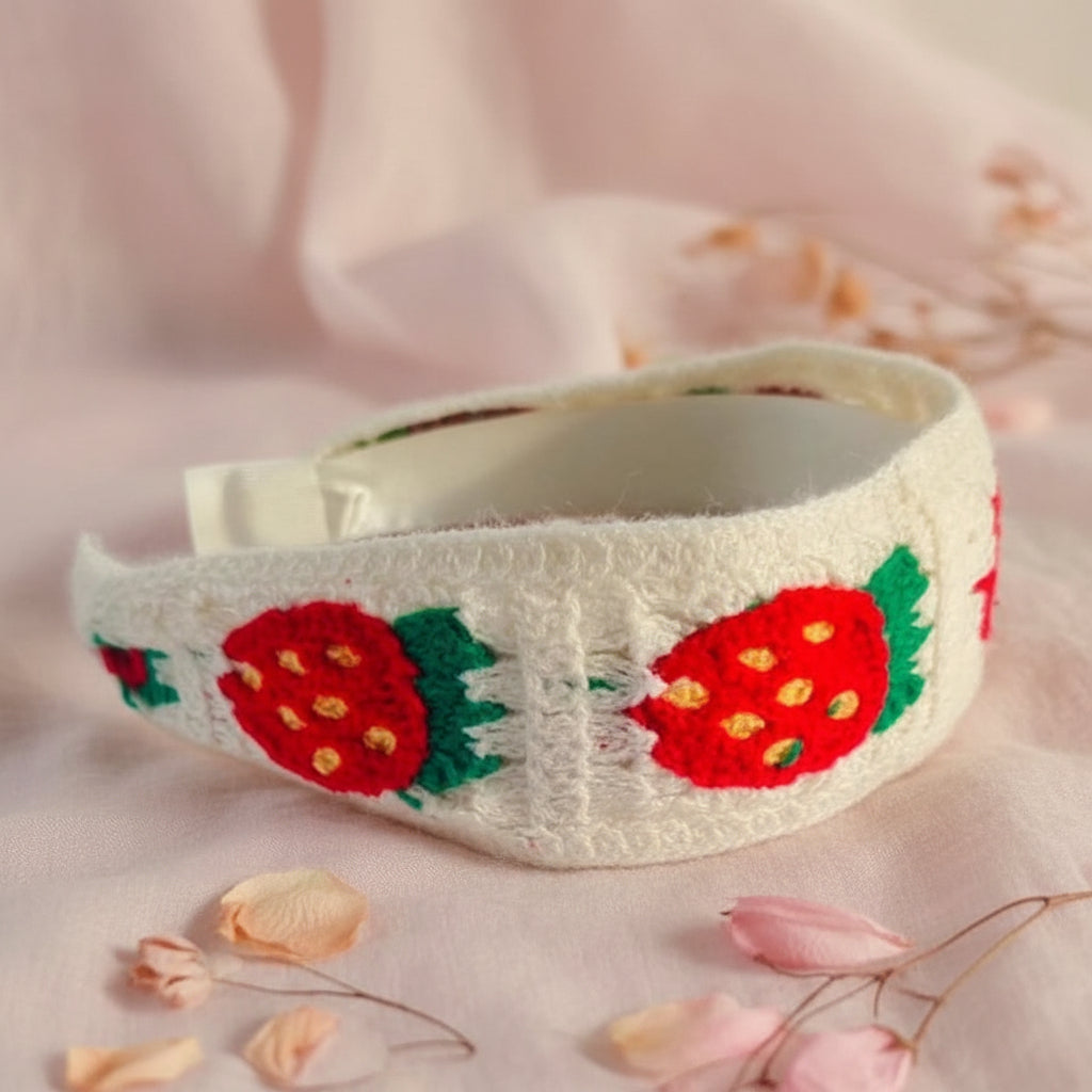 A white textured fabric headband featuring embroidered red strawberries with green leaves and yellow centers, displayed on a white faux fur surface.