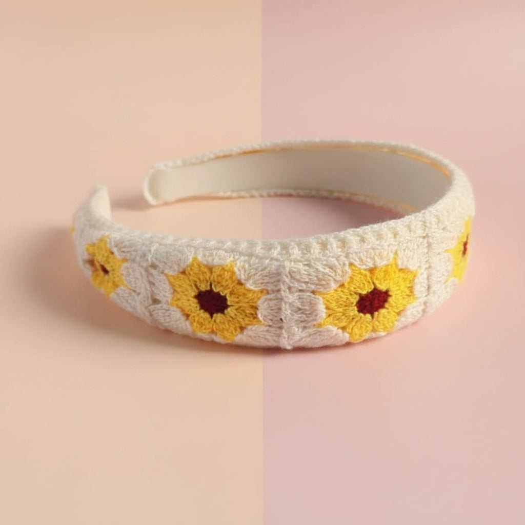 A white crochet headband decorated with yellow sunflowers with red centers, resting on a white faux fur surface.